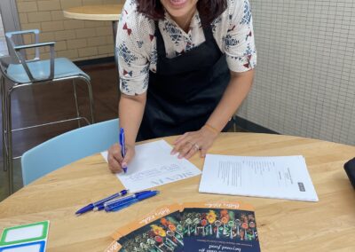 A woman smiling at a table with papers, pens, during the Lebanese Cooking Class with Jihan